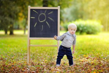 Beautiful toddler boy drawing standing by a blackboard outdoors